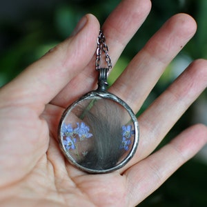 Könnte beinhalten: Eine runde, silberfarbene Anhängerkette mit einem durchsichtigen Glasmittelteil. Das Glas zeigt eine kleine Anordnung getrockneter blauer Blumen und dunkles Haar. Der Anhänger hängt an einer filigranen Kette.