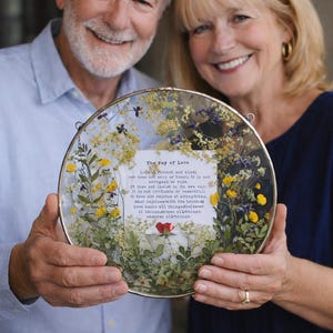 May include: A circular framed artwork with dried flowers and text reading "The Fay of Love." The artwork is held by two people. The frame has a gold-coloured edge. The artwork includes yellow, purple, and green elements.