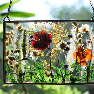 May include: A rectangular glass panel with a pressed flower arrangement. It features a red rose, orange lily, and dried flowers and greenery, encased in clear resin. The panel is framed with dark metal and suspended by a chain. The overall effect is a botanical display.