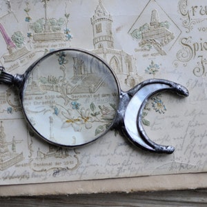 Puede incluir: Colgante de lupa plateado con un asa en forma de media luna. La lente redonda magnifica una postal vintage con ilustraciones florales y arquitectónicas. El colgante cuelga de una delicada cadena.