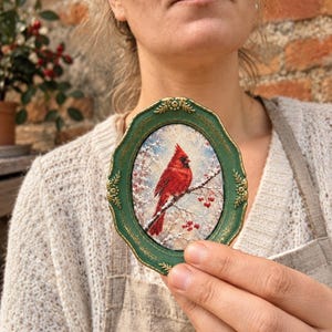 May include: An oval-shaped decorative frame in a dark green finish with gold-toned accents, containing an embroidered fabric piece that depicts a bright red cardinal bird perched on a snowy branch with small red berries against a winter background.