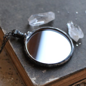 May include: A silver-toned, round, hand-held mirror with a chain attached. The mirror is on a dark gray surface with two clear quartz crystals.