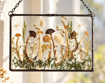 Puede incluir: Un colgante de pared rectangular de vidrio enmarcado en metal oscuro con una cadena para colgar. El interior contiene setas secas prensadas, musgo verde, paniculata blanca, hierbas altas y cabezas de semillas blancas y esponjosas.