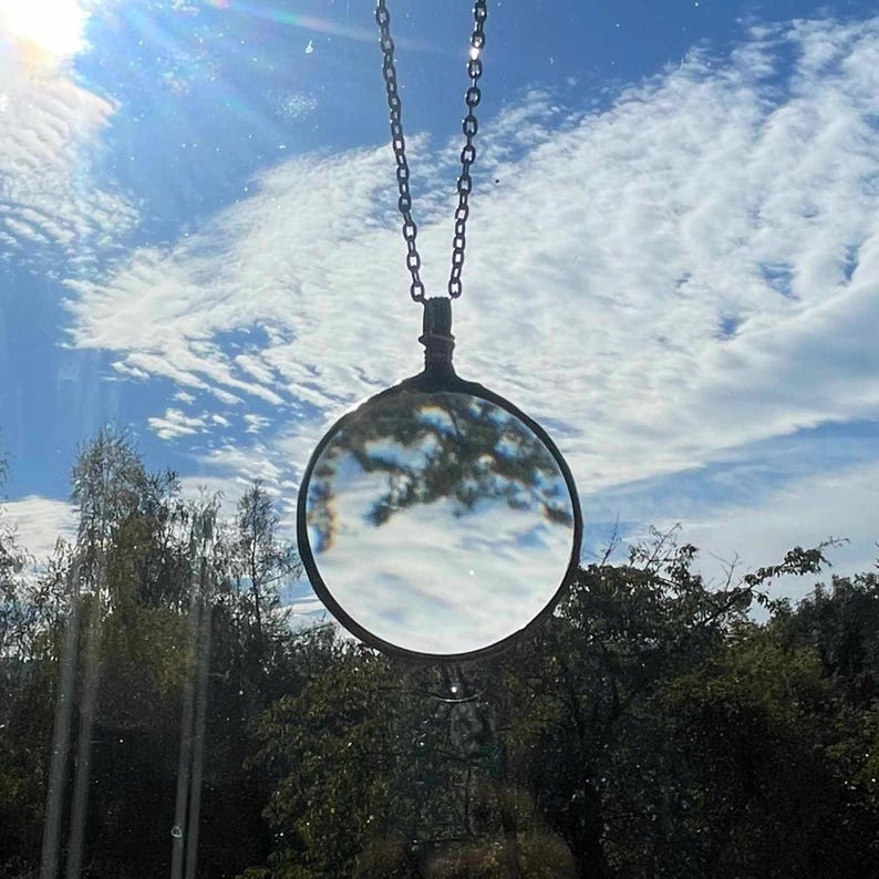 K&ouml;nnte beinhalten: Eine silberne Kettenkette mit einem runden Lupenanh&auml;nger. Das Glas reflektiert einen blauen Himmel mit wei&szlig;en Wolken.