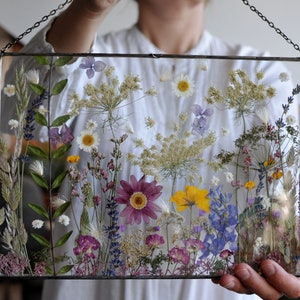 Könnte beinhalten: Ein rechteckiger Glasrahmen mit einer Vielzahl von gepressten Blumen in Lila, Gelb, Weiß und Blau. Die Blumen sind so angeordnet, dass sie eine natürliche, botanische Darstellung bilden, die in dem klaren Glas schwebt.
