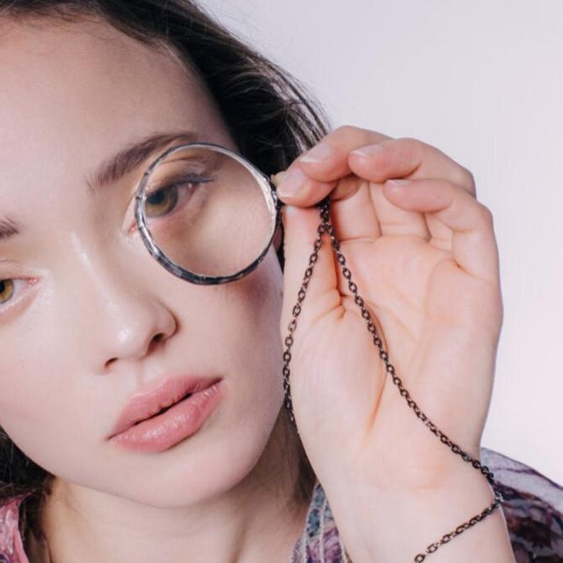 May include: A silver-toned monocle with a chain, held up to the eye. The monocle has a circular lens and a decorative frame. The chain is dark and delicate, adding a touch of elegance. The background is a soft, neutral color.
