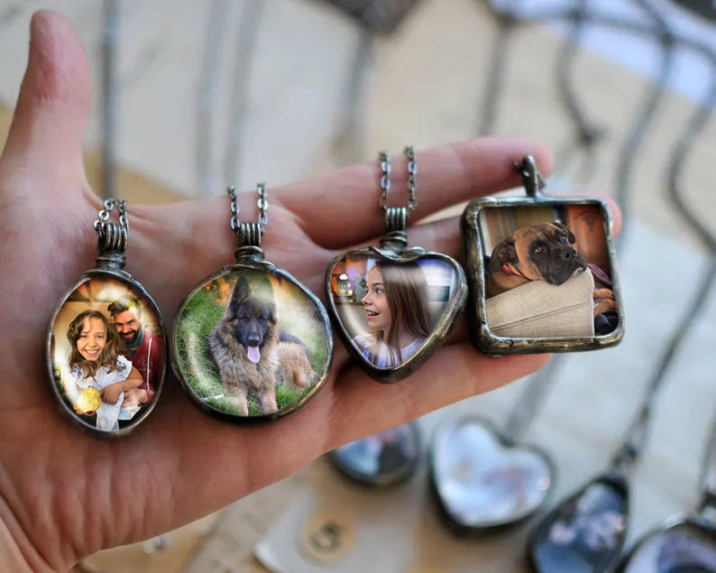 May include: Four silver necklaces with glass pendants featuring different photos. The pendants are round, heart-shaped, and square. The photos include a family, a dog, and a girl.
