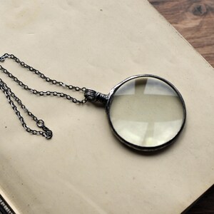 May include: A silver-toned magnifying glass pendant necklace with a round lens. The magnifying glass is attached to a delicate chain. The pendant rests on a light brown surface, possibly a book page.