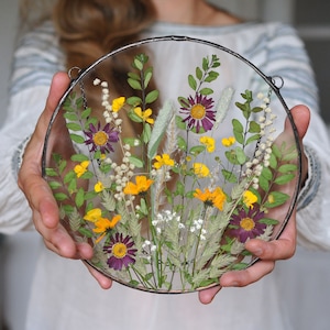 May include: A round glass frame with a pressed flower arrangement. The flowers are a mix of purple, yellow, and white. The frame is hanging from a chain.