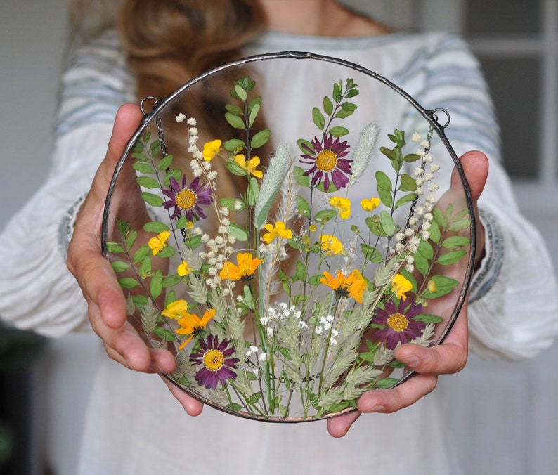May include: A round glass frame with a pressed flower arrangement. The flowers are a mix of purple, yellow, and white. The frame is hanging from a chain.