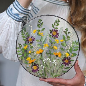 May include: A round glass frame with a pressed flower arrangement. The frame is suspended from a silver chain and features a variety of yellow, purple, and white flowers.