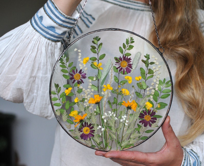 May include: A round glass frame with a pressed flower arrangement. The frame is suspended from a silver chain and features a variety of yellow, purple, and white flowers.