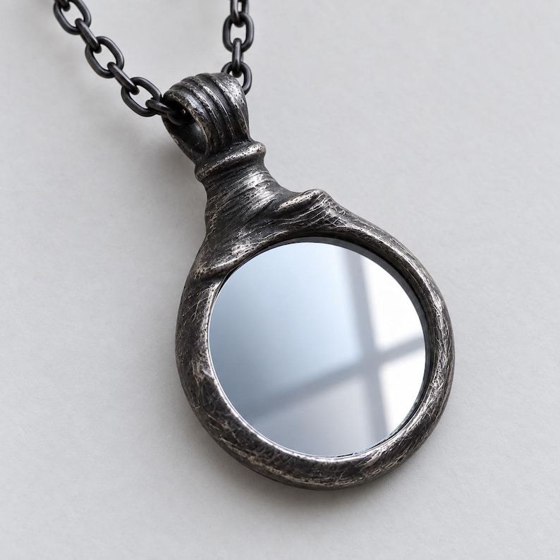 May include: A circular pendant necklace featuring a dark, weathered, metallic frame with a textured, distressed finish. The center of the pendant contains a smooth, reflective mirror surface. The piece hangs from a matching dark metal chain link.