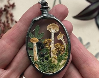Puede incluir: Un colgante de forma ovalada con un marco de metal oscuro y texturizado que contiene elementos botánicos preservados. Dentro de la carcasa transparente hay tres setas de color claro, follaje verde, pequeñas flores amarillas y bayas oscuras.