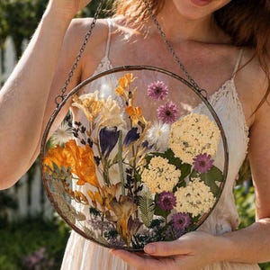 Op de afbeelding: Een ronde, heldere glazen bloemenkunst met een metalen frame en ketting. Het kunstwerk toont gedroogde oranje, gele, paarse en witte bloemen. Het stuk wordt opgehangen door een zilveren ketting.