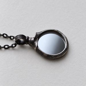 May include: A pendant necklace featuring a circular, dark, reflective stone set within a textured, oxidized silver frame. The pendant is attached to a dark metal chain with oval links, showcasing a weathered, metallic finish throughout.