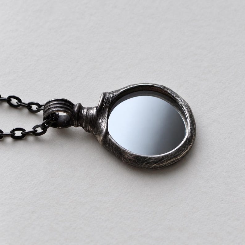 May include: A pendant necklace featuring a circular, dark, reflective stone set within a textured, oxidized silver frame. The pendant is attached to a dark metal chain with oval links, showcasing a weathered, metallic finish throughout.