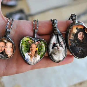 May include: Four silver necklaces with glass pendants featuring different images. The pendants are round, heart-shaped, and oval. One pendant shows two girls, another shows a girl in a garden, another shows a cat, and the last shows a girl with a black cat.