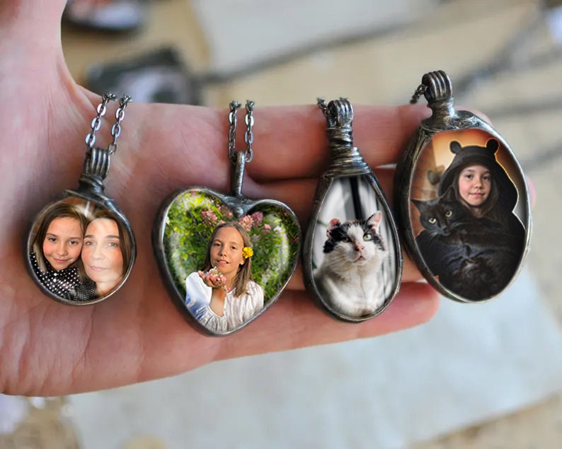 May include: Four silver necklaces with glass pendants featuring different images. The pendants are round, heart-shaped, and oval. One pendant shows two girls, another shows a girl in a garden, another shows a cat, and the last shows a girl with a black cat.