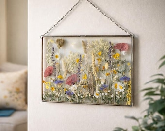 May include: A rectangular framed pressed flower art piece. The artwork features a variety of dried flowers in shades of red, yellow, white, and purple. The frame is a dark metal with a chain for hanging. The artwork is displayed against a white wall.