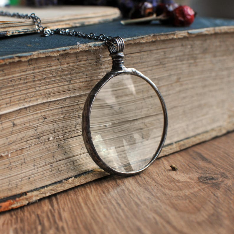 May include: A silver-toned magnifying glass pendant with a clear lens hangs from a delicate chain. The pendant rests on an open, aged book with worn pages. The background features a wooden surface and dried flowers, creating a vintage aesthetic.