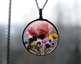 May include: A silver chain necklace with a round pendant featuring pressed flowers in shades of pink, yellow, and purple. The flowers are encased in clear resin.