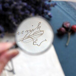 May include: A clear magnifying glass focuses on a handwritten signature in gray ink on white paper. The background includes dried berries and a blue surface, with a hint of purple flowers. The image is a close-up, highlighting the details.