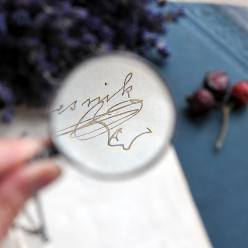 May include: A clear magnifying glass focuses on a handwritten signature in gray ink on white paper. The background includes dried berries and a blue surface, with a hint of purple flowers. The image is a close-up, highlighting the details.