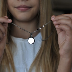 May include: A silver-toned necklace featuring a circular pendant with a smooth, reflective, white center. The pendant is suspended from a delicate, matching silver-toned metal chain with small, uniform oval links that connect to the top bail.