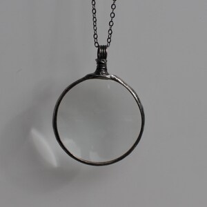 May include: A silver-toned magnifying glass pendant with a clear lens. The round lens is framed by a dark metal border and hangs from a matching chain. The pendant has a vintage or antique aesthetic.