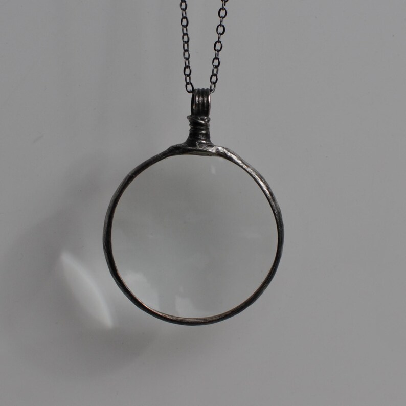 May include: A silver-toned magnifying glass pendant with a clear lens. The round lens is framed by a dark metal border and hangs from a matching chain. The pendant has a vintage or antique aesthetic.