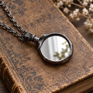 May include: A circular pendant necklace featuring a reflective mirror surface encased in a dark, weathered, textured metal frame with a matching link chain. The pendant has a rugged, aged appearance with visible grooves and a metallic finish.