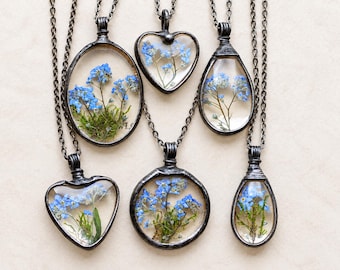 Peut inclure: Six colliers avec des cadres en métal sombre contenant de la résine transparente avec des fleurs myosotis bleues pressées et de la mousse verte. Les pendentifs sont ovales, en forme de cœur, circulaires et en goutte, sur des chaînes assorties.
