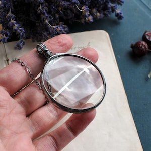 May include: A silver-toned magnifying glass pendant with a clear lens and a delicate chain. The round pendant is held in a hand, with a vintage paper and dried lavender in the background. The pendant's lens is approximately 2 inches in diameter.