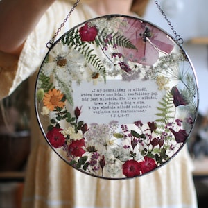 Pode incluir: Um ornamento redondo de vidro com uma corrente prateada para pendurar. O ornamento está cheio de flores e vegetação prensadas, incluindo samambaias, rosas e flores silvestres. Um cartão branco com uma citação escrita à mão em polonês é visível no centro do ornamento.