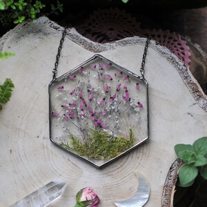 May include: A hexagonal glass pendant necklace with a pressed flower design. The pendant features a cluster of pink and white flowers with green moss at the base. The necklace is made with a silver chain.