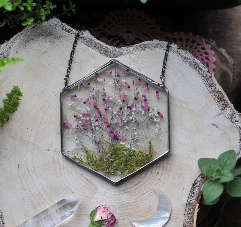 May include: A hexagonal glass pendant necklace with a pressed flower design. The pendant features a cluster of pink and white flowers with green moss at the base. The necklace is made with a silver chain.