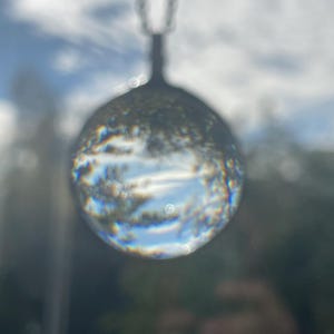 K&ouml;nnte beinhalten: Eine Halskette mit einem Anh&auml;nger aus klarem Glas in Kugelform und einer silbernen Kette. Die Kugel reflektiert den Himmel und die Wolken und erzeugt einen vertr&auml;umten und &auml;therischen Effekt.