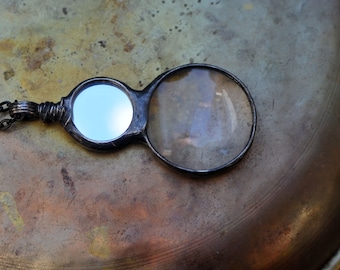 May include: A silver-toned magnifying glass necklace with a round magnifying lens and a smaller round mirror. The necklace has a chain.