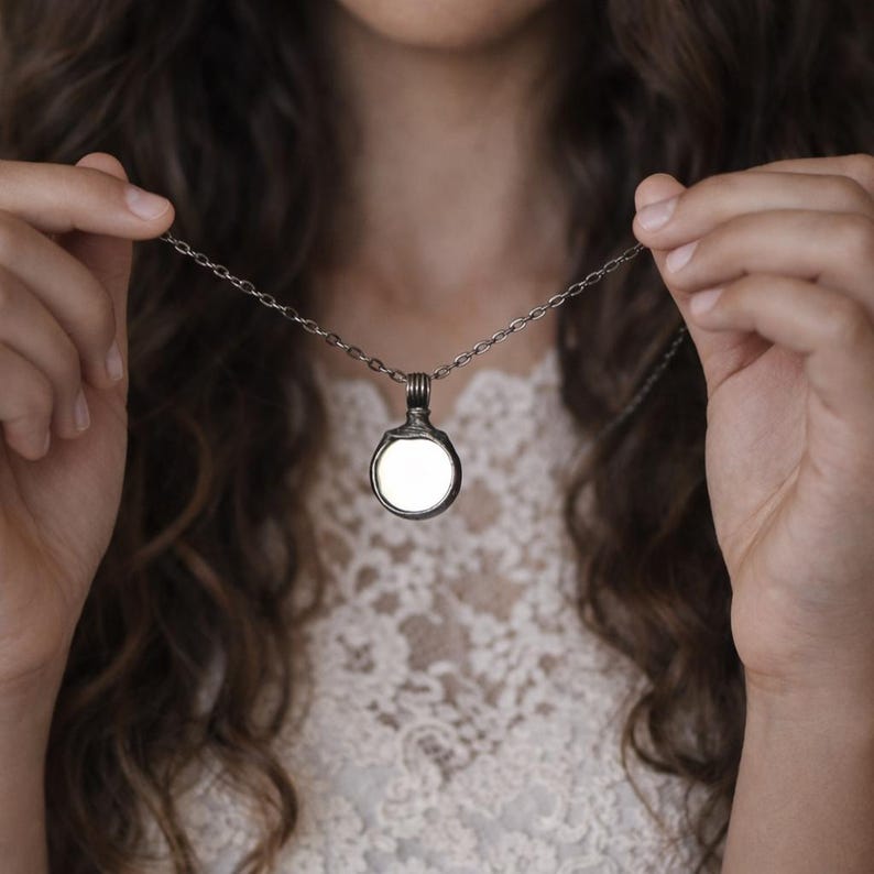 May include: A circular pendant necklace featuring a dark, metallic, textured frame surrounding a smooth, white, reflective center. The pendant hangs from a matching dark metal chain with small, uniform, oval-shaped links.