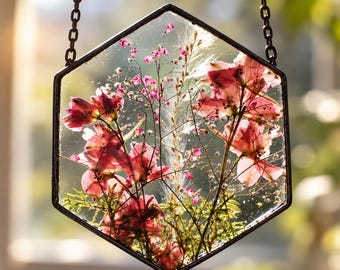 May include: A hexagonal glass wall hanging featuring pressed pink flowers, green foliage, and delicate white grasses encased between two clear panes. The piece is framed in a thin black metal border and suspended by a matching black metal chain.