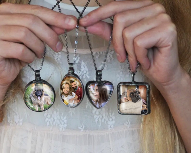 May include: Four silver necklaces with glass pendants featuring different photos. The pendants are round, oval, heart-shaped, and square. The photos include a dog, a family, a girl, and a dog.