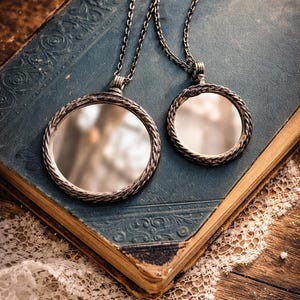 May include: Two silver-toned pendant necklaces with round, mirrored faces and braided bezels rest on a vintage blue book. The book has ornate embossed designs and aged, yellowed pages. A delicate white lace doily is draped nearby.