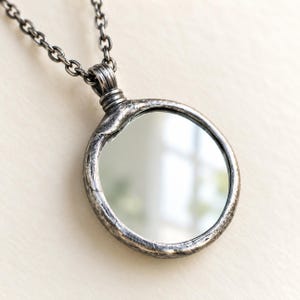 May include: A circular pendant necklace featuring a reflective mirror surface encased in a textured, dark oxidized silver frame. The pendant hangs from a matching dark silver chain with a series of interconnected oval links, creating a cohesive look.