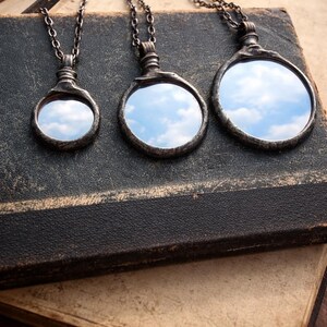 Peut inclure: Trois colliers pendentifs de style antique avec des faces rondes en miroir reflétant un ciel nuageux. Les pendentifs sont sertis dans des cadres en métal foncé ornés et sont suspendus à de fines chaînes. Les colliers reposent sur un livre vintage à la couverture usée et texturée.