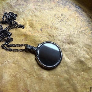 May include: A dark metallic pendant featuring a circular, reflective black surface encased in a textured, oxidized frame. The pendant is attached to a matching dark metal chain with elongated, interconnected links that extend outward.