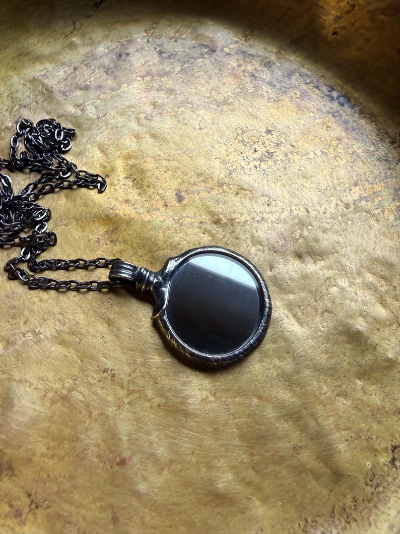 May include: A dark metallic pendant featuring a circular, reflective black surface encased in a textured, oxidized frame. The pendant is attached to a matching dark metal chain with elongated, interconnected links that extend outward.