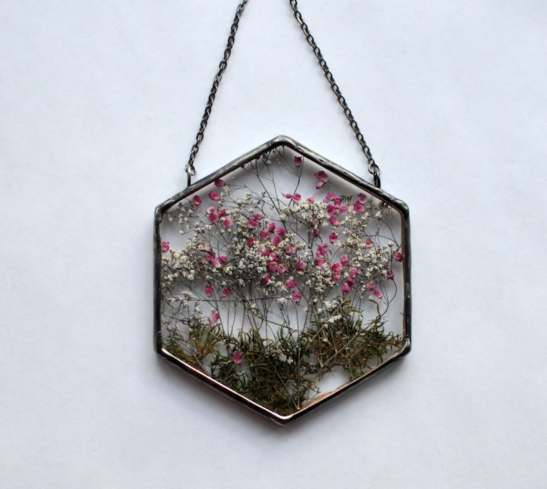 May include: A hexagonal glass ornament with a silver metal frame and chain. The ornament contains pressed dried flowers and greenery, including pink flowers and green leaves.