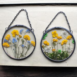 May include: Two circular glass art pieces with pressed flowers. One has yellow and blue flowers, the other yellow and white flowers with green moss. Each has a silver chain for hanging.