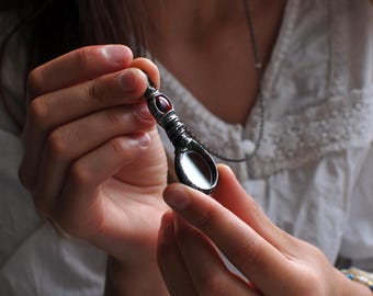 May include: A silver-toned pendant necklace with a small, oval mirror and a red gemstone. The pendant is held by a person. The pendant has a detailed, textured design around the mirror and gemstone.